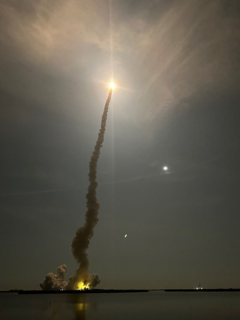 NASA's Artemis I rocket mid-launch. A bright circle of light ascends with a trail of gray plumed of smoke.