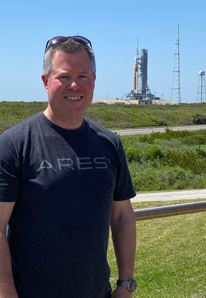 John Youskauskas smiles in the foreground wearing a T-shirt that reads ARES. Behind him is NASA's SLS rocket, green shrubbery and a blue sky.