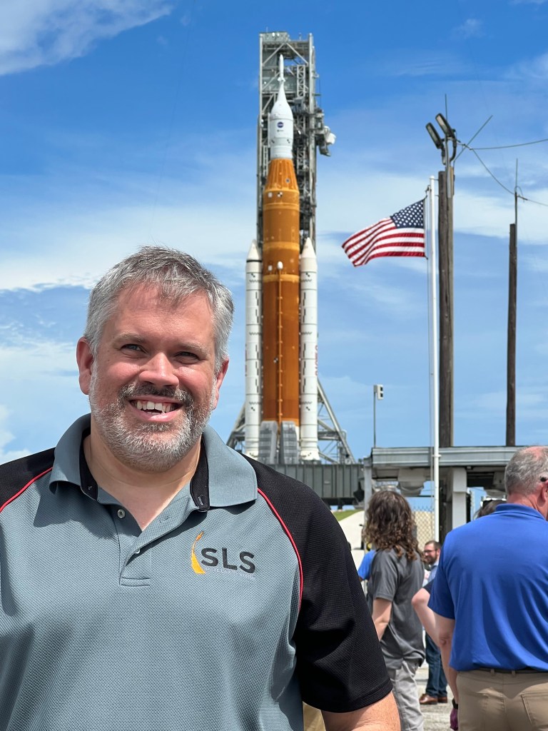 David Hitt is smiling in the foreground, behind him is NASA's SLS rocket, a crowd of onlookers, and a clear blue sky.