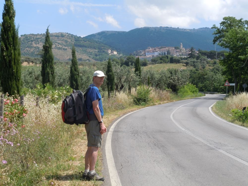 On a sunny day, Bill Thorness stands beside a paved road in Rome. Behind him are foliage-covered mountains.