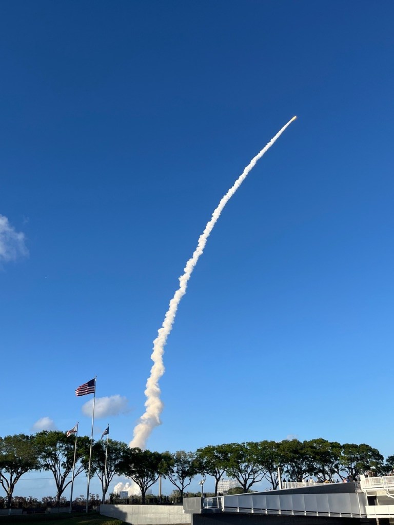 Artemis II launch, a plume of smoke streaks into the blue sky, in the foreground are trees and the American flag.