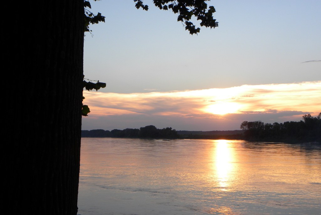 The sun sets on the horizon beyond horizontal clouds. The sunset is reflected in the muddy waters of the Missouri.