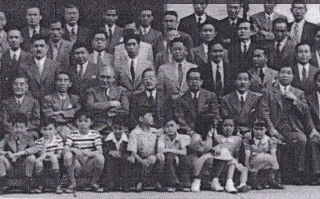 Black and white still from recovered footage of Japanese diplomats; they stand posed in rows with their children in front.