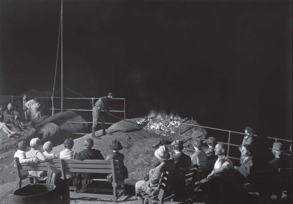 People seated on wooden benches watch as a man pushes glowing embers off a cliff in the dark.